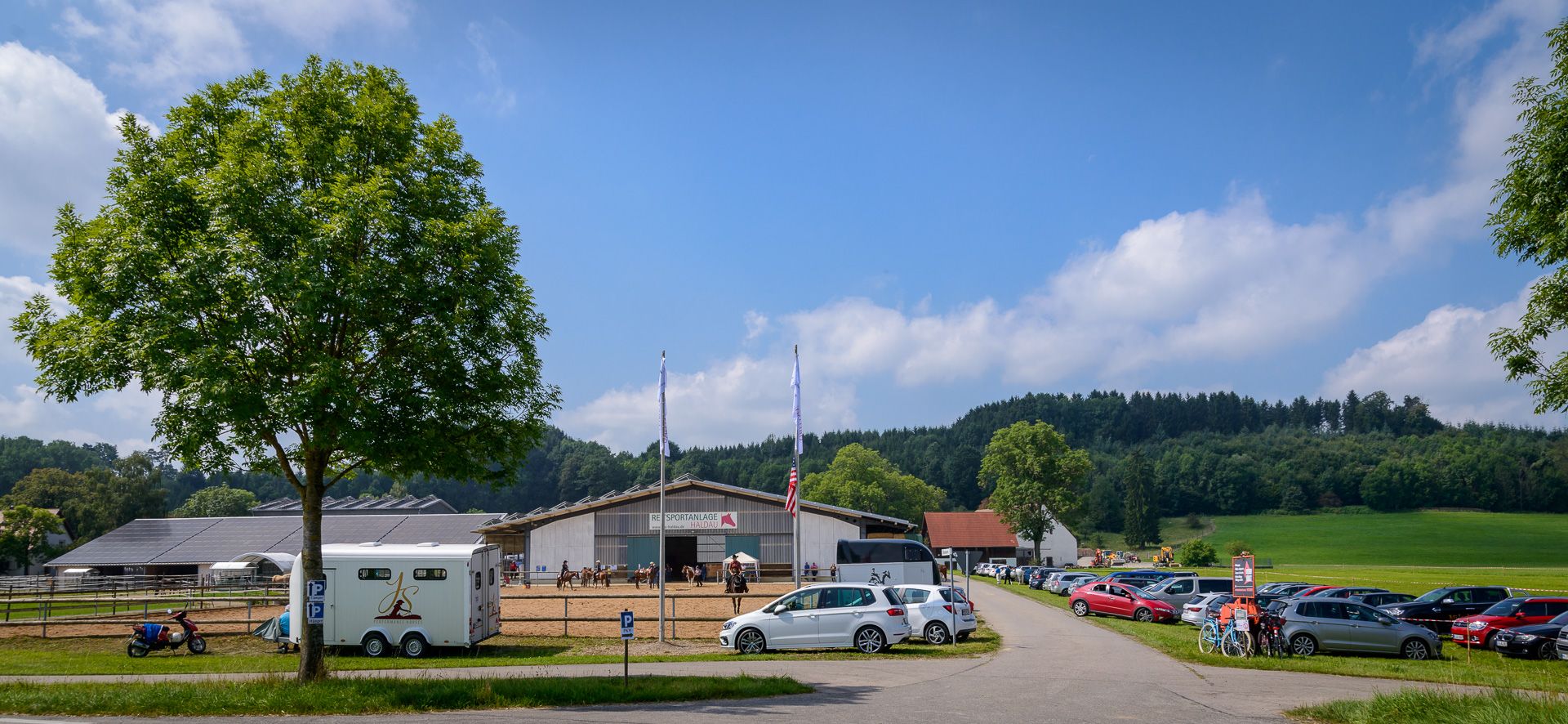 Zufahrt zur Reitsportanlage Haldau mit Parkplatz, Pferdeanhänger und Reithalle bei Sonne
