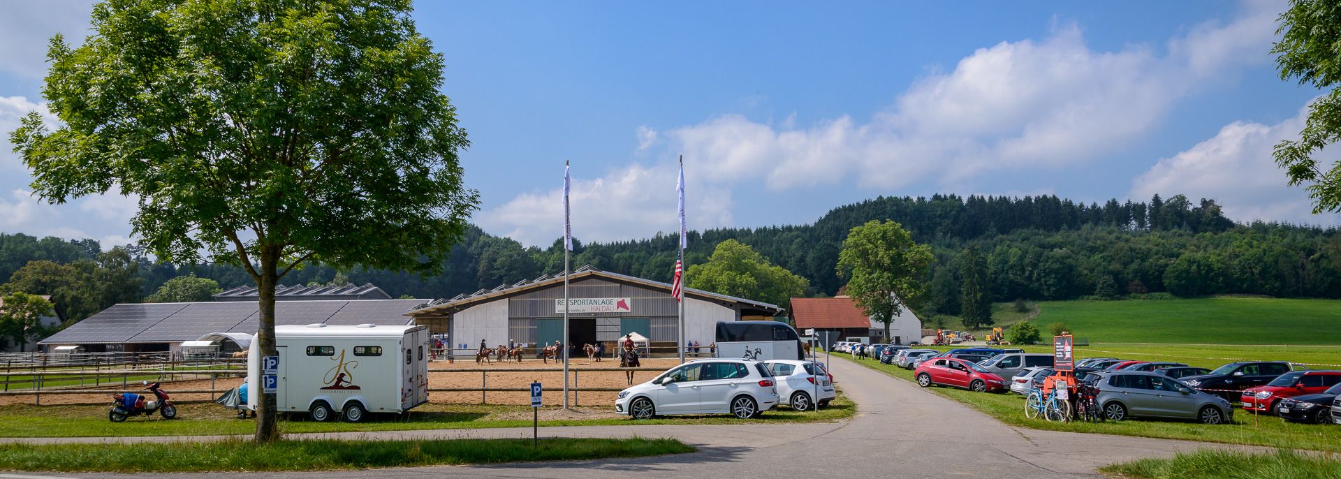 Zufahrt zur Reitsportanlage Haldau mit Parkplatz, Pferdeanhänger und Reithalle bei Sonne
