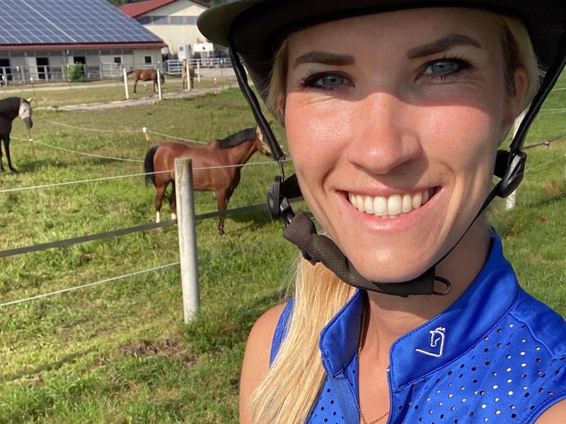Melanie Müller mit Reithelm, im Hintergrund Pferde auf der Koppel und der Stall
