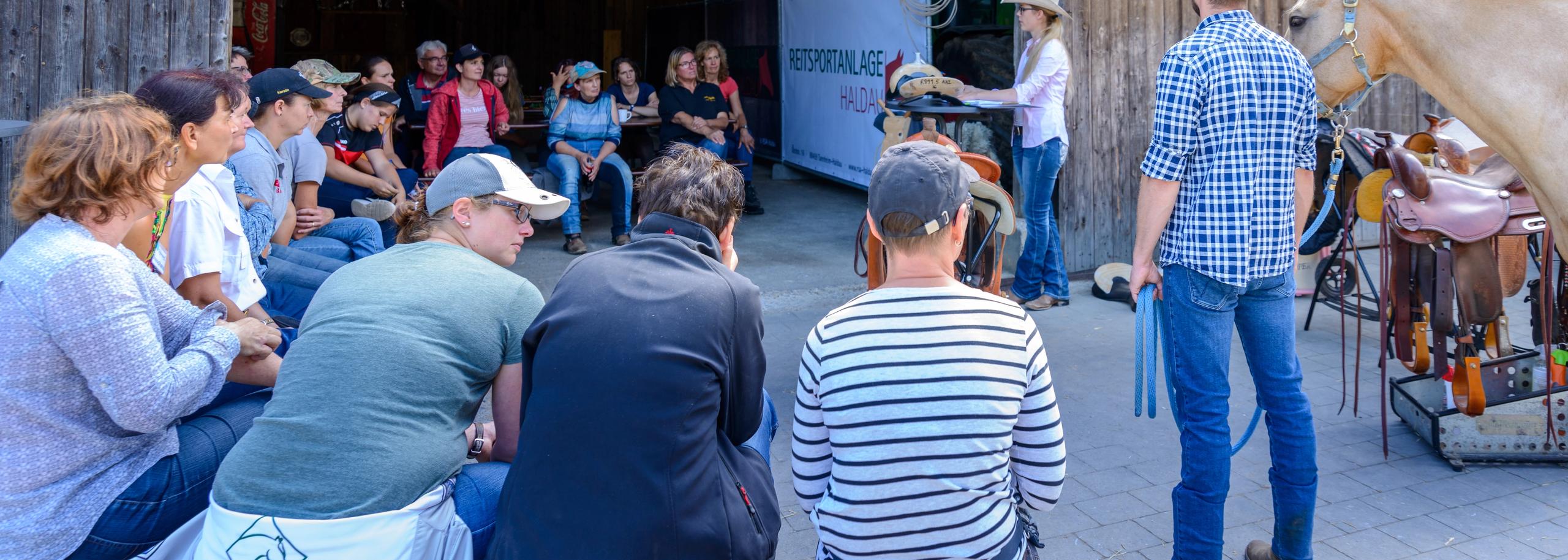 Trainer erklärt einer Gruppe Reiterinnen und Reiter vor der Scheune die Arbeit am Pferd